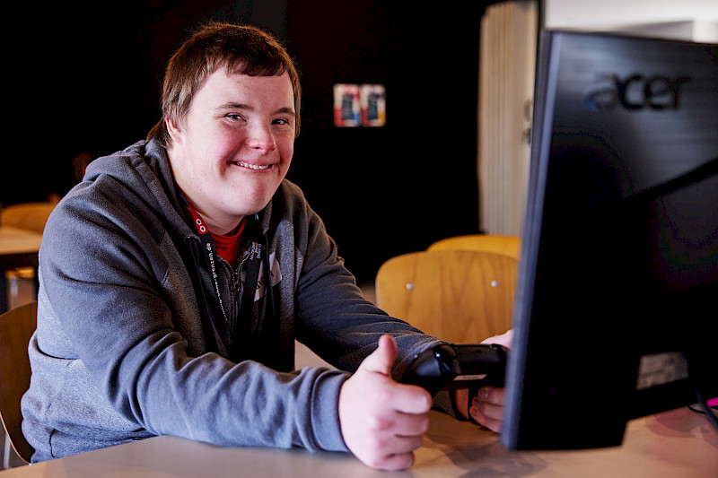 Ein Junge spiel mit einem Controller vor einem Bildschirm. Er schaut in die Kamera, lächelt und zeigt einen Daumen nach oben.