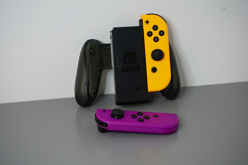 Ein Bild von einer Joy-Con-Halterung. Nur ein Joy-Con ist eingesteckt. Der Zweite liegt vor der Halterung.