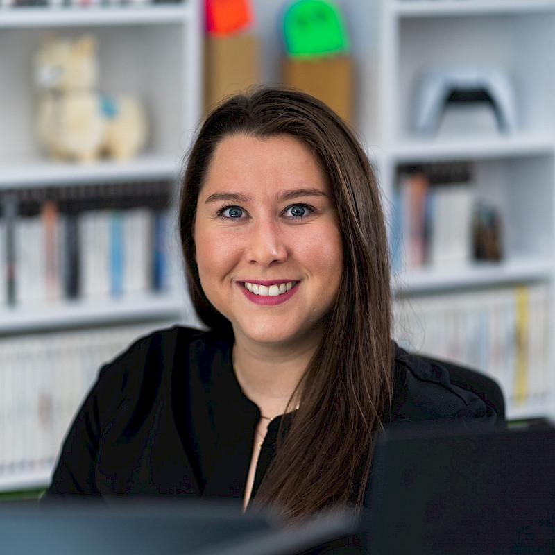 Saskia hat lange braune Haare. Sie sitzt vor einem PC. Hinter ihr ist ein Regal mit Spielen.
