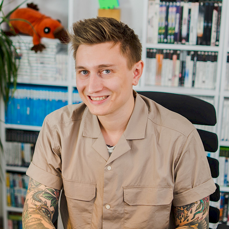 Julius hat kurze, blonde Haare und trägt ein beiges Hemd. Er sitzt vor einem Regal mit Spielen.