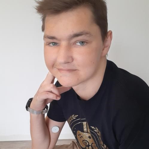 Dennis stützt seinen Kopf auf der Hand ab. Er hat kurze, blonde Haare und trägt ein schwarzes Tshirt. Er lächelt.