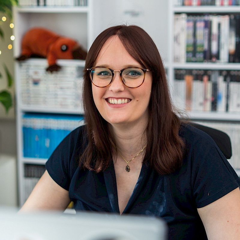 Karolina hat lange braune Haare. Sie trägt eine Brille und ein dunkles Oberteil. Sie sitzt vor einem Regal mit Spielen.