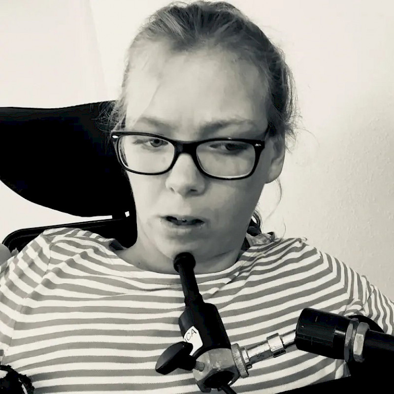 Marlene hat helle Haare und trägt eine Brille. Das Bild ist schwarz-weiß. Sie sitzt im Rollstuhl und nutzt einen Kinn-Joystick.