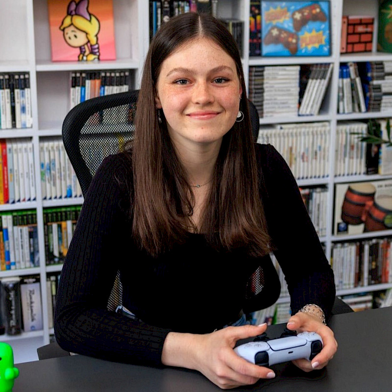 Lisa hat einen Controller in der Hand. Sitzt an einem Schreibtisch und lächelt in die Kamera.