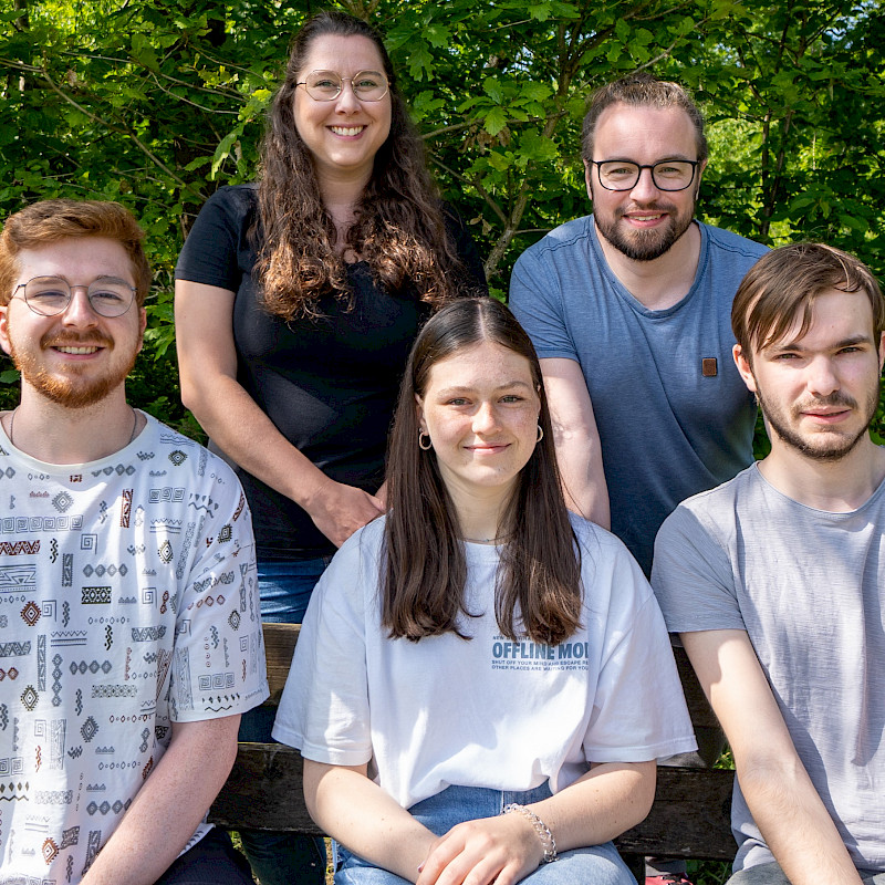 Levi, Lisa und Martin sitzen auf einer Bank. Saskia und Sven stehen dahinter. Sie sind vor einer grünen Hecke im Hintergrund.