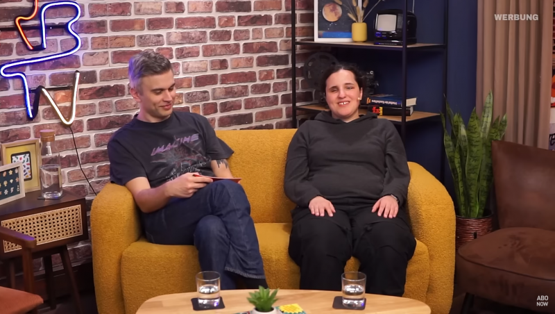 Fabian und Svenja sitzen gemeinsam auf einer gelben Couch. Fabian hat kurze graue Haare. Svenja hat lange braune Haare zu einem Zopf zusammengebunden.
