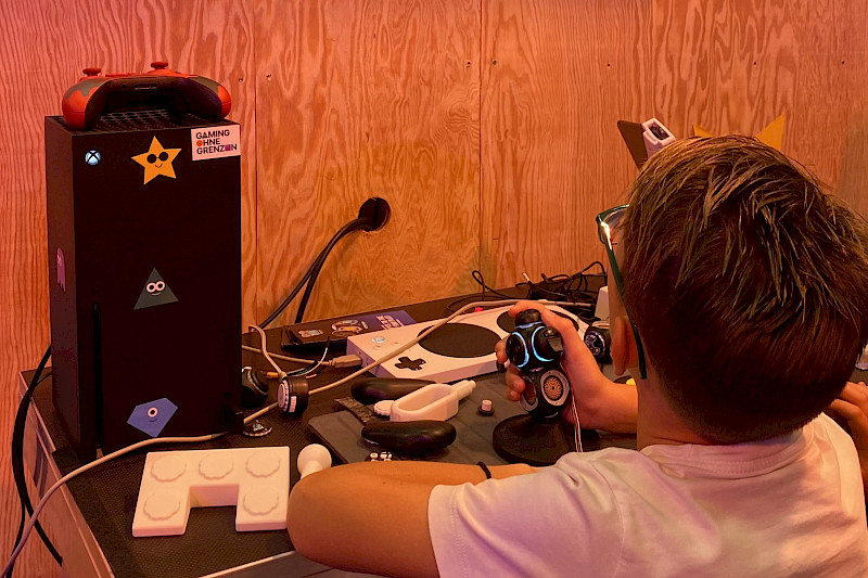 Ein Junge spielt Forza Horizon 5 mit einem Proteus Controller. Der Controller ist ein schwarzer Joystick mit Knöpfen. Er schaut auf einem Bildschirm vor ihn auf der Wand.