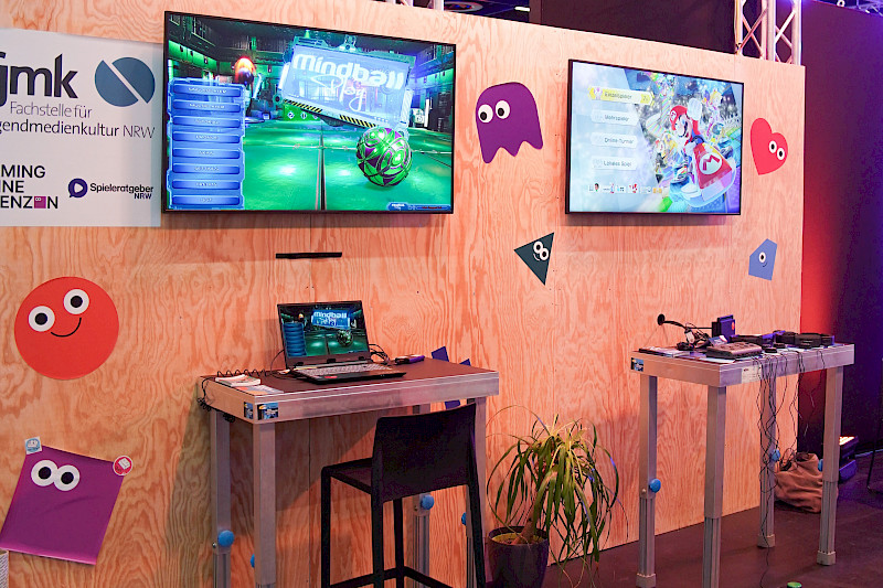 Der Stand auf der Messe. Zwei höhere Tische auf denen ein Laptop mit einem Eyetracker und eine Switch mit einem Hori Flex Controller stehen. Darüber jeweils ein Monitor. Mit dem Eyetracker wird MIndball gespielt. Mit der Switch Mario Kart.