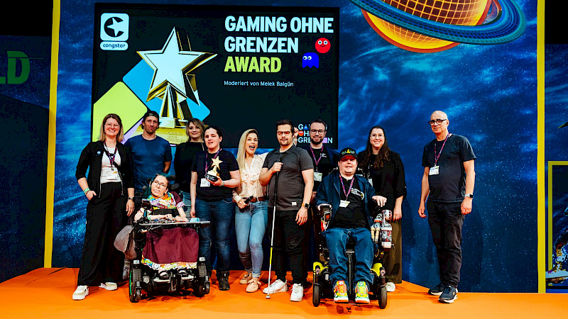 Alle Beteiligten vom Award sind gemeinsam auf der Bühne für ein Gruppenfoto. Im Hintergrund steht Gaming ohne Grenzen Award auf einer LED Leinwand. Es ist ein großer goldener Pokal in Sternform noch mit abgebildet.