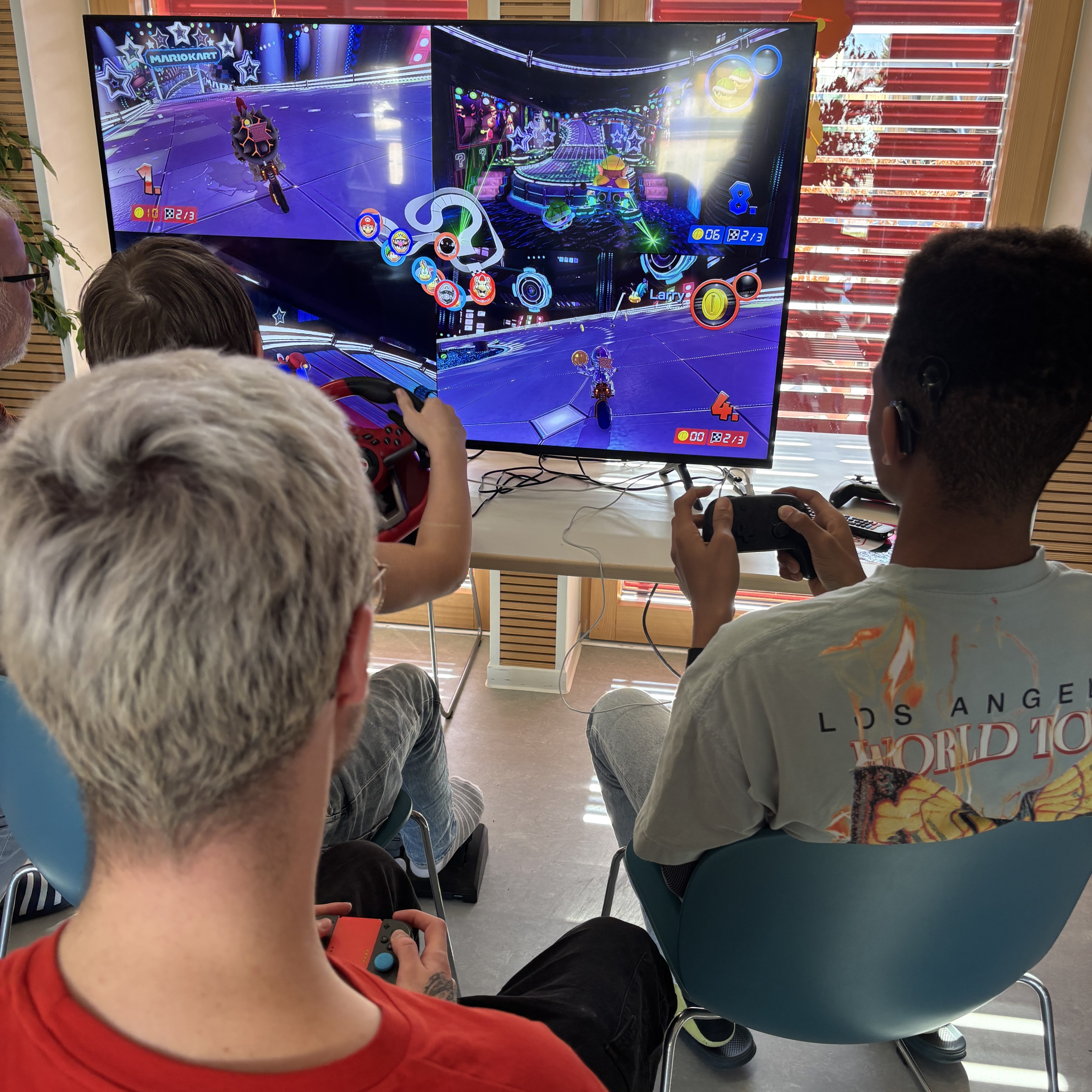 Drei Jugendliche sitzen vor einem Fernseher. Sie haben alle einen Controller in der Hand. Einer spielt an einem Lenkrad. Sie spiele Mario Kart.