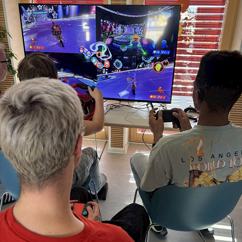 Drei Jugendliche sitzen vor einem Fernseher. Sie haben alle einen Controller in der Hand. Einer spielt an einem Lenkrad. Sie spiele Mario Kart.