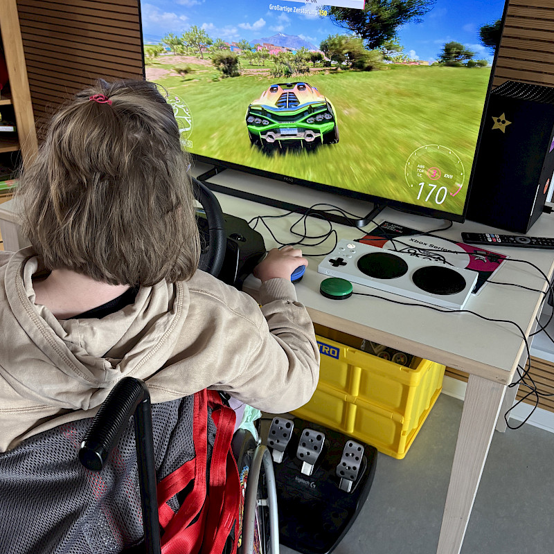Eine Jugendliche mit längeren Haaren sitzt im Rollstuhl. Sie steuert ein Autorennspiel mit einem Lenkrad und dem Adaptive Controller. Sie schaut auf einen Fernseher.