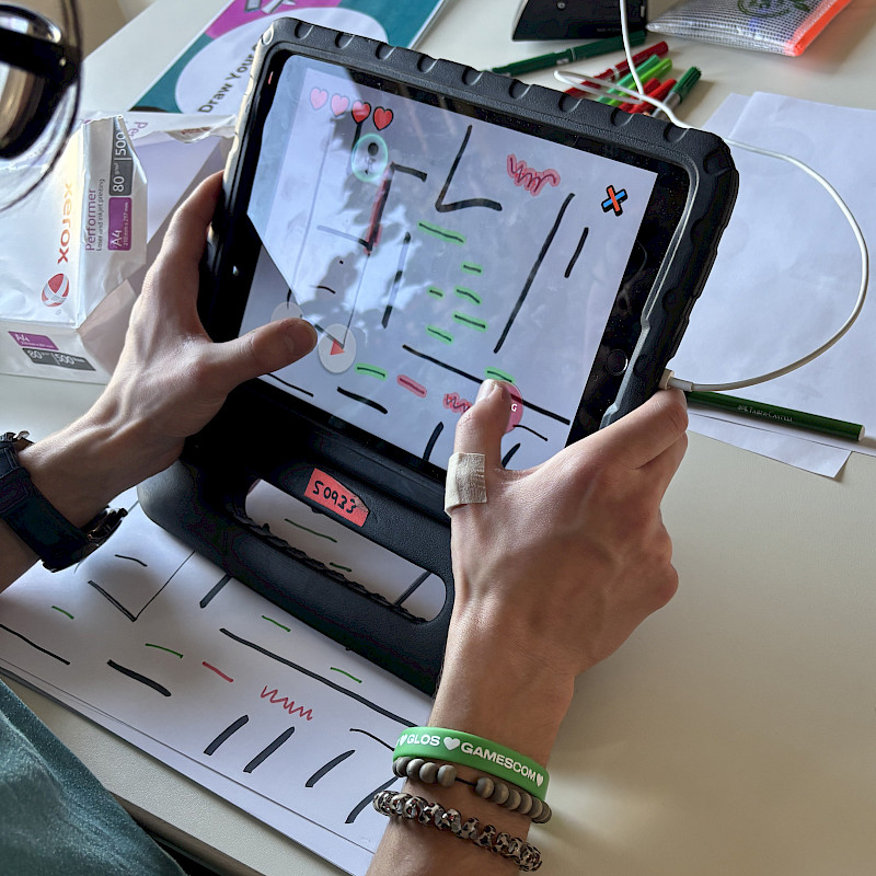 Eine Person hält ein iPad in der Hand. Darauf ist die App Draw Your Game offen. Ein gemaltes Spiel kann auf dem iPad digital gespielt werden.