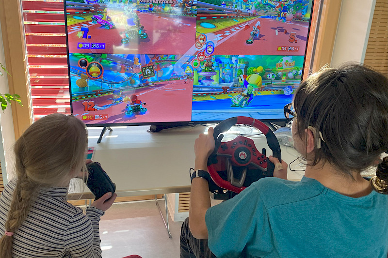 Zwei Jugendliche mit langen Haaren sitzen vor einem Fernseher. Eine Person spielt mit einem Controller. Eine Person spielt an einem Lenkrad. Sie spielen Mario Kart.