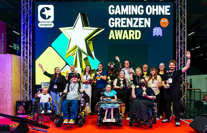 Ein Foto vom Gaming ohne Grenzen Award 2025. Alle Laudator*innen und Preisträger*innen sind auf einer Bühne. Sie lachen und manche winken in die Kamera. Im Hintergrund ist ein Bild projiziert. Auf dem Bild ist die Trophäe und das Logo von congstar.