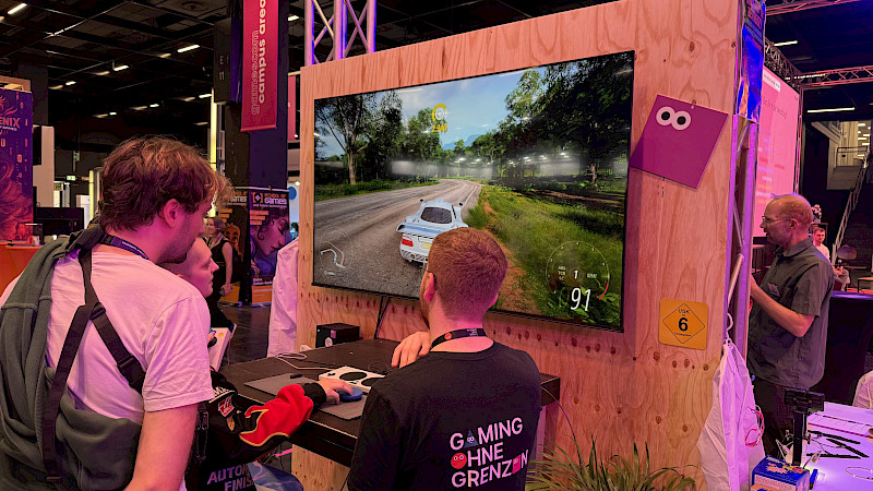 Auf dem Foto sind drei Personen. Sie stehen oder sitzen vor einem großen Bildschirm. Auf dem Bildschirm fährt ein Auto. Die Personen spielen an der Station.