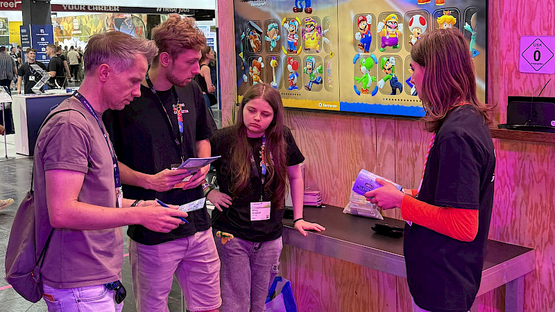 Auf dem Foto sind vier Personen vor der Mario Kart World Station. Drei Jugendliche aus Level Up erklären einem Mann das Spiel.