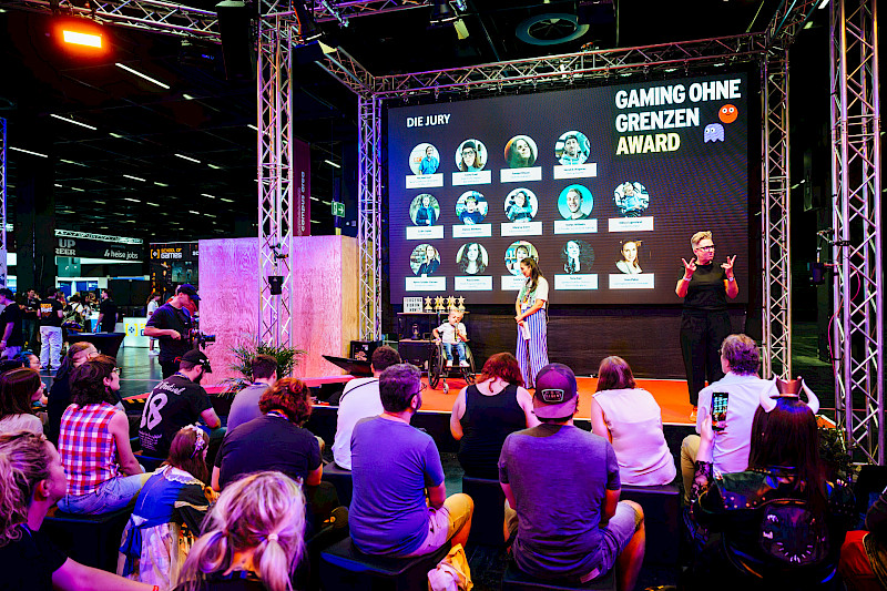 Menschen auf einer Bühne. Ein Publikum vor der Bühne. Eine Gebärdensprachdolmetscherin übersetzt. Auf einer großen Leinwand steht Gaming ohne Grenzen Award und die Personen aus der Jury sind abgebildet.