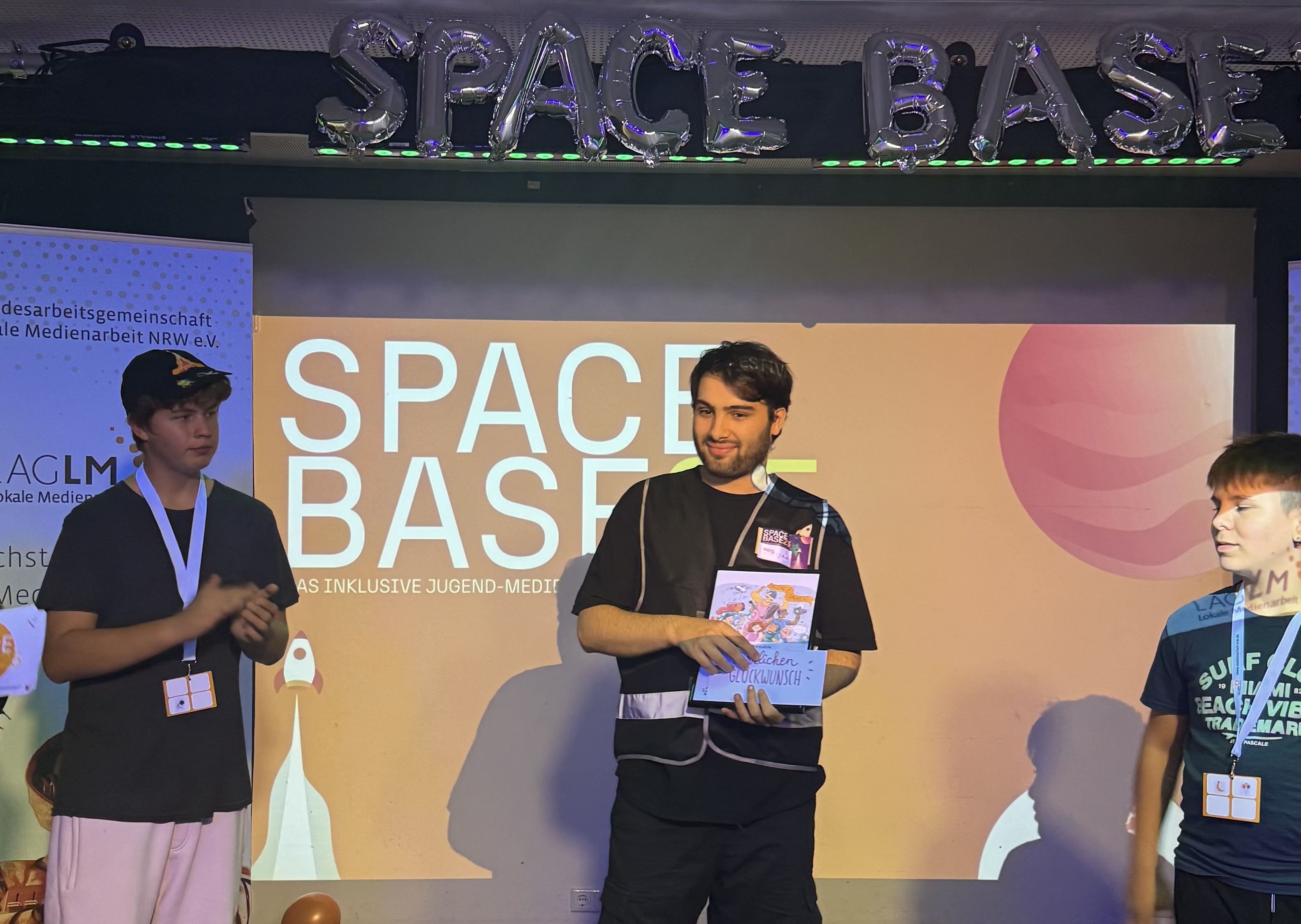 Drei Jugendliche stehen auf einer Bühne. Sie halten Preise in der Hand. Auf der Leinwand hinter ihnen steht: SpaceBase 25.