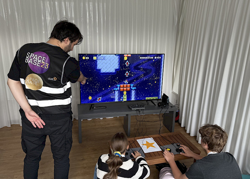 2 Jugendliche spielen ein Spiel. Eine Person steht daneben und erklärt etwas.