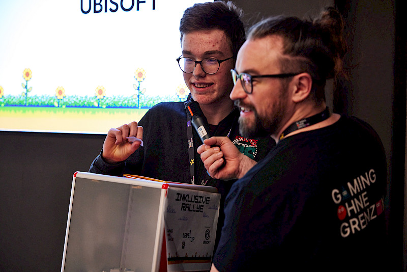 Zwei Menschen stehen nebeneinander vor einem Bildschirm. Eine Person hält ein Mikrofon in der Hand. Die andere Person zeigt auf etwas vor sich. Vor ihnen steht eine Box mit der Aufschrift „Inklusive Rallye“. Im Hintergrund ist ein Bildschirm mit einem Spi