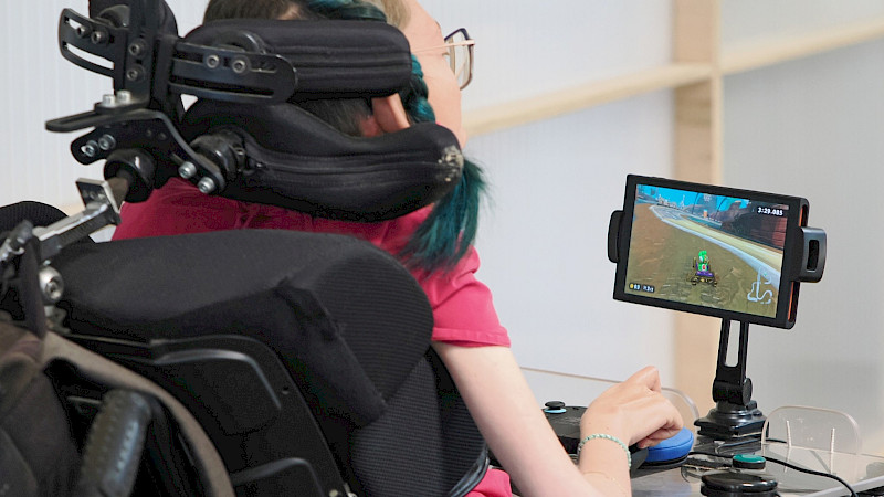 Eine Person sitzt in einem Rollstuhl mit Kopfstütze und spielt ein Videospiel auf einem kleinen Bildschirm. Sie nutzt angepasste Eingabegeräte auf einem Tisch vor sich, während auf dem Bildschirm ein Rennspiel zu sehen ist.