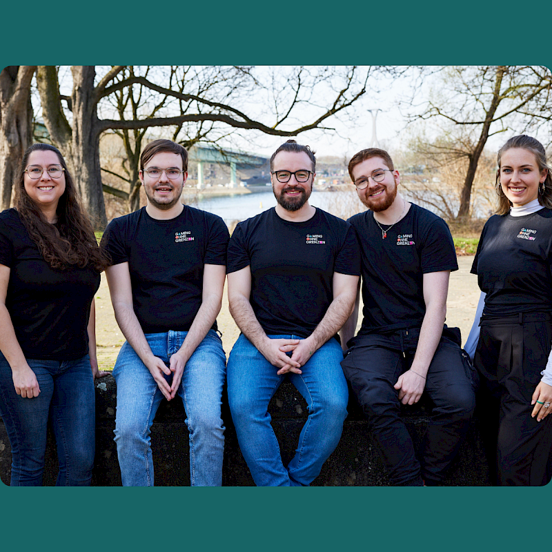 Fünf junge Erwachsene tragen schwarze T-Shirts mit Logo und posieren gemeinsam im Freien. Drei sitzen auf einer niedrigen Steinmauer, zwei stehen daneben. Im Hintergrund sind ein Fluss, eine Brücke und kahle Bäume zu sehen. Alle schauen in die Kamera.
