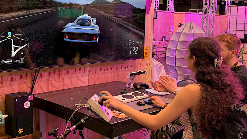 Zwei Personen spielen an einem Messestand gemeinsam ein Rennspiel mit dem adaptive Controller.