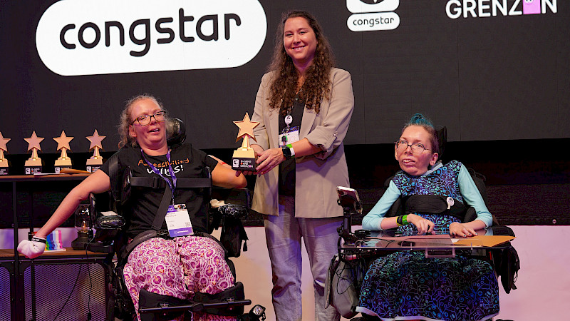 Zwei Frauen im Rollstuhl und eine stehende Frau präsentieren gemeinsam eine goldene Stern-Trophäe beim „Gaming ohne Grenzen Award“. Im Hintergrund sind weitere Trophäen sowie das Congstar- und Veranstaltungslogo zu sehen.